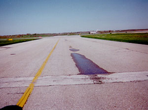 Overview photo of a taxiway pavement with visible low-severity rutting in one wheel path.           The rutting is visible because there is standing water in the wheel path.