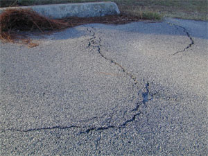 Close-up photo showing an upward bulge in the pavement surface near the pavement's edge.           The upward bulge is very noticeable due to a crack in the pavement surface at the apex of the bulge.